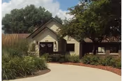 Front entrance of a single-story beige rehabilitation and care center building with a curved driveway and landscaped bushes and trees.