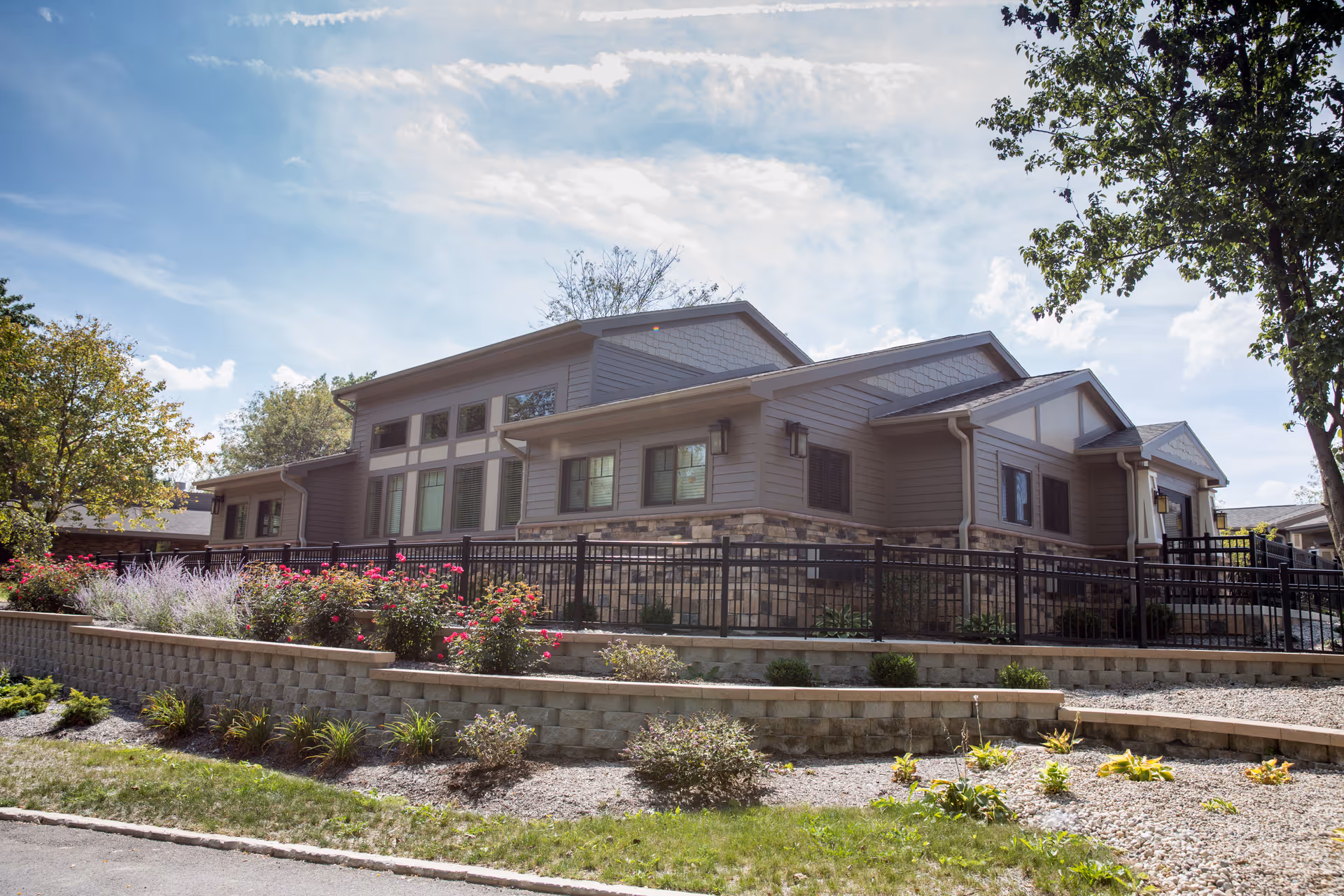 Front exterior of a single-story senior living building with terraced stone planters, flowering shrubs, and a black metal fence under a partly cloudy sky.