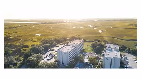 Aerial view of two multi-story residential buildings surrounded by parking lots and expansive marshland with the sun low on the horizon.