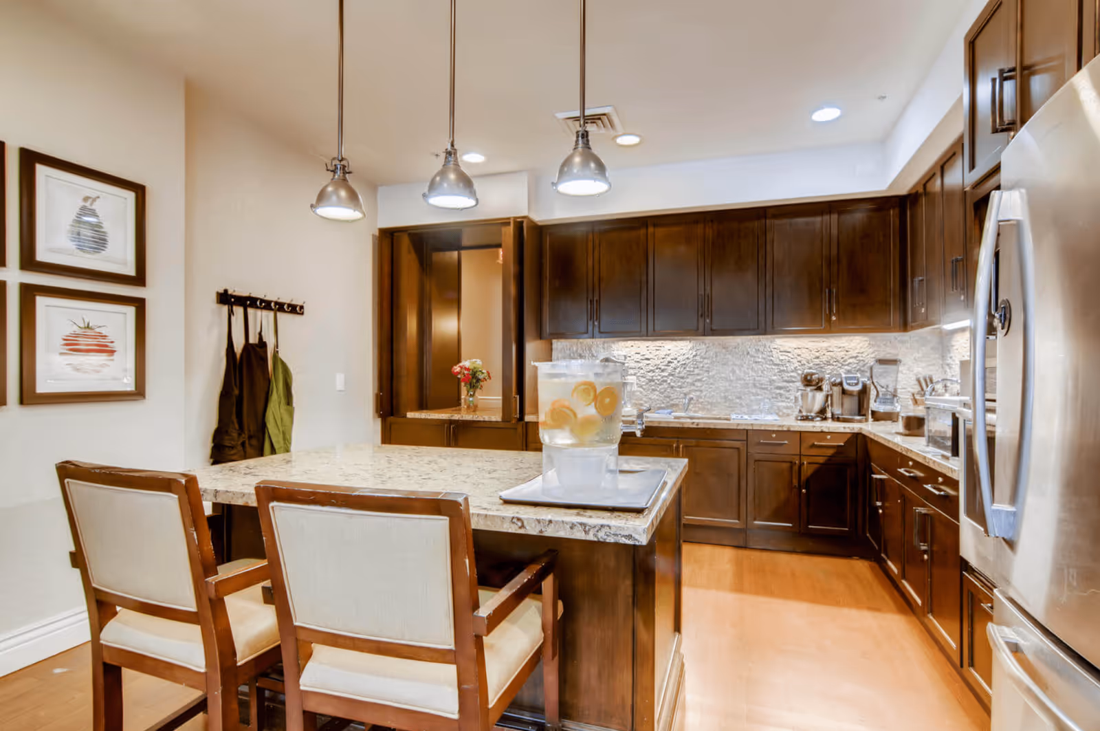 A modern kitchen with dark wooden cabinets, a light granite countertop island with two cushioned chairs, three hanging pendant lights, and stainless steel appliances. A water dispenser with lemon slices is placed on the island. There are two framed fruit artworks on the wall and aprons hanging on hooks.