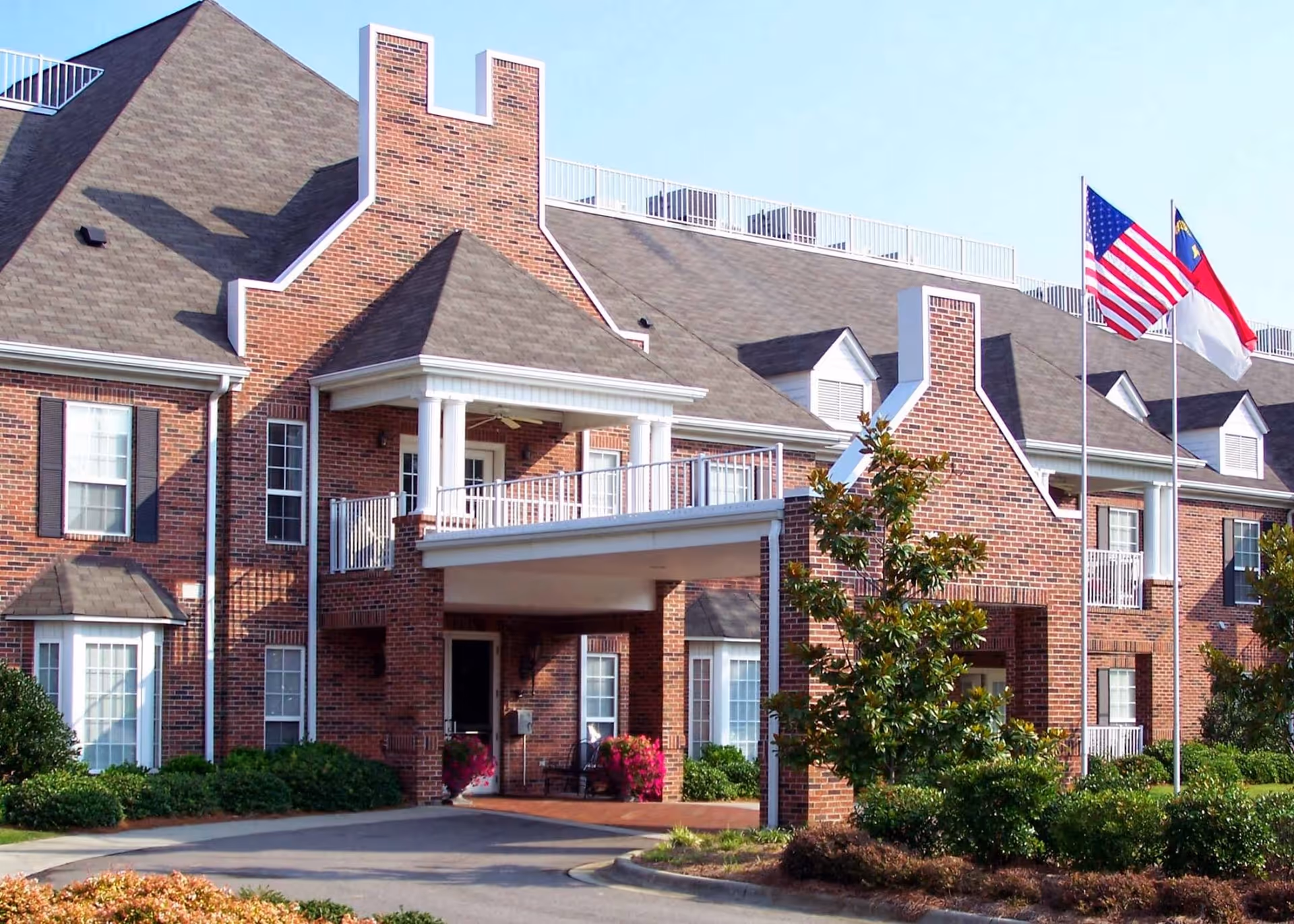 Exterior view of a brick senior living facility building with multiple windows, a covered entrance with white columns, and two flagpoles displaying the American and North Carolina state flags. The building is surrounded by well-maintained landscaping including bushes and small trees.