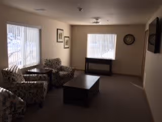 A small living room area with two patterned armchairs, a wooden coffee table, and a console table beneath a window with vertical blinds. The walls are decorated with framed pictures and a clock. The room has carpeted flooring and neutral-colored walls.