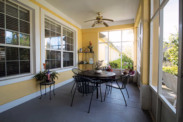 A screened-in porch area with a round metal table and four matching chairs. The porch has yellow walls, two large windows, a ceiling fan with lights, and several potted plants on a metal shelf and small tables. The porch overlooks a garden area outside.
