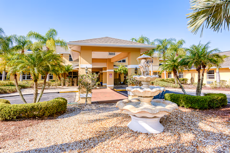 Front exterior view of Sun City Senior Living facility with a three-tiered decorative fountain in the foreground, surrounded by gravel and landscaping with palm trees and bushes. The building has a covered entrance with the number 3855 displayed above.