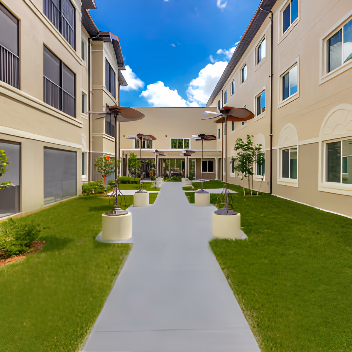 Outdoor courtyard area between two beige multi-story buildings with windows, green grass lawns on either side of a central concrete walkway, and several modern metal sculptures mounted on cylindrical bases along the path under a blue sky with scattered clouds.