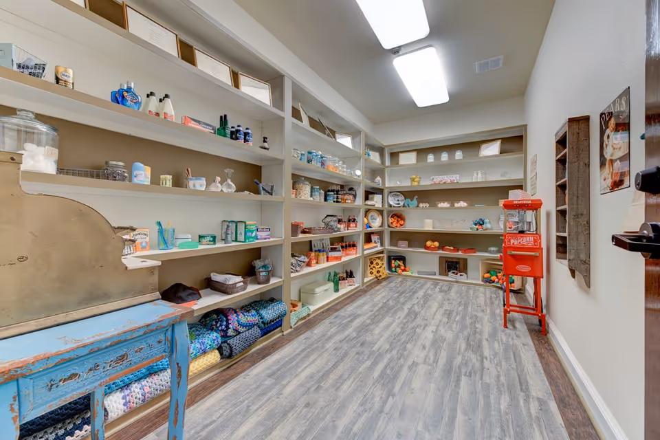 Interior view of a small community shop/pantry with wall shelves stocked with toiletries, snacks, decor and a red popcorn machine.