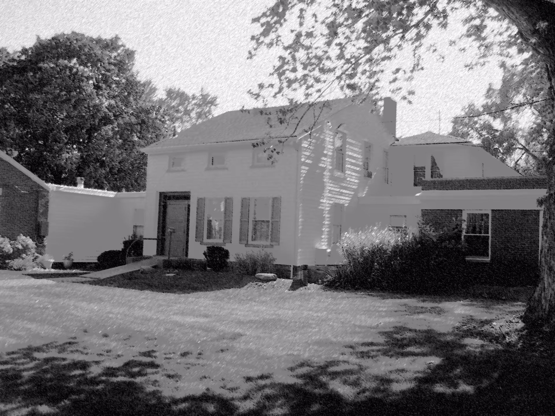 Exterior view of a two-story residential building with white siding and multiple windows, surrounded by trees and bushes, with a grassy lawn in the foreground.