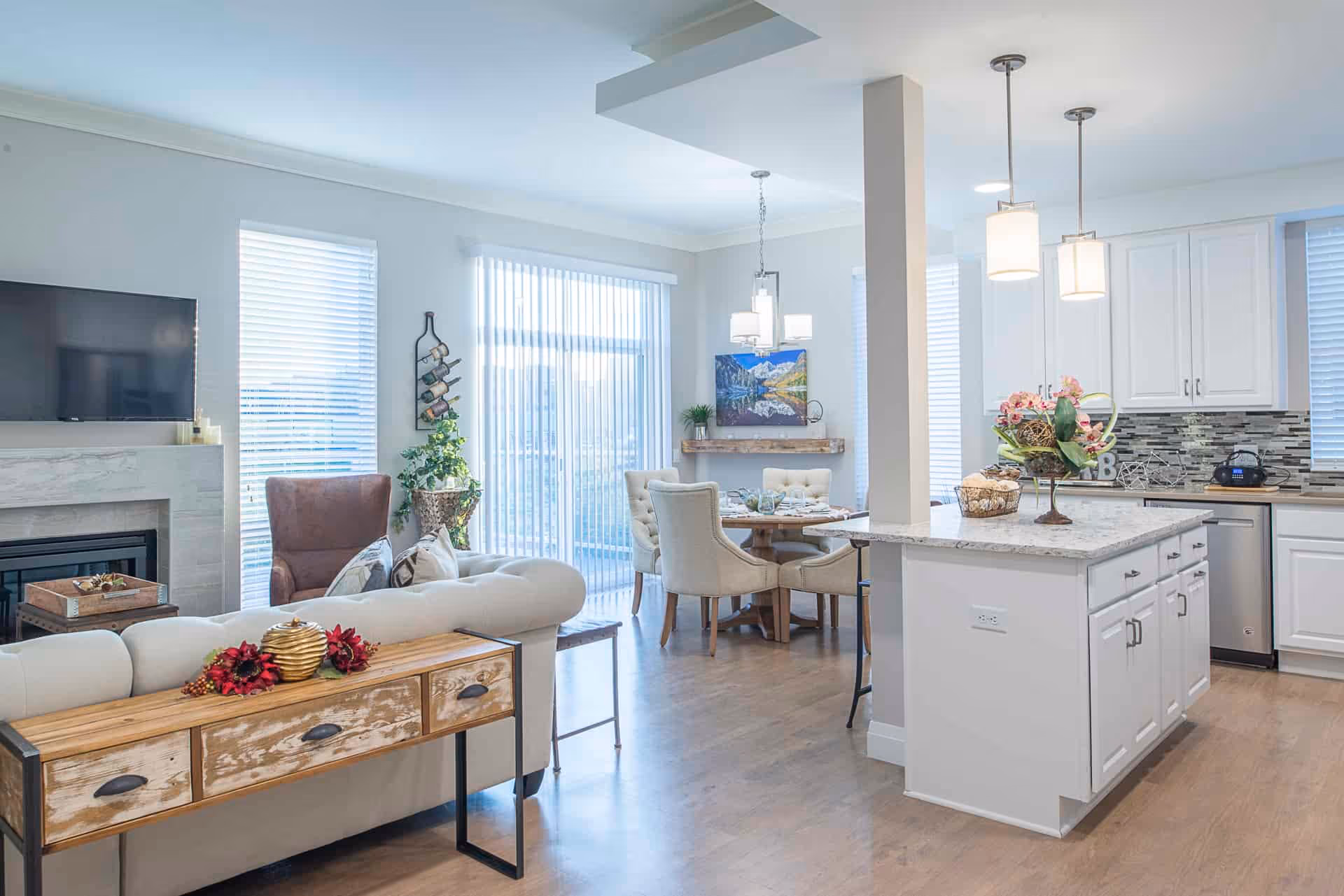 Bright and modern open-concept living area featuring a white sofa with decorative pillows, a wooden console table with fall-themed decorations, a brown armchair, a fireplace with a mounted flat-screen TV, a dining area with a round table and upholstered chairs, and a kitchen with white cabinets, a marble island, pendant lights, and stainless steel appliances.