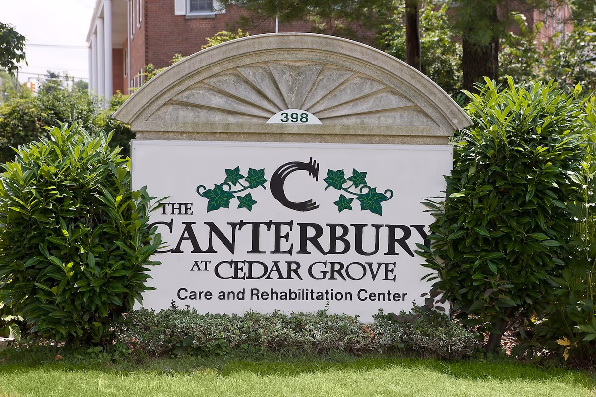 Outdoor sign for The Canterbury at Cedar Grove Care and Rehabilitation Center surrounded by green bushes and grass, with part of a brick building visible in the background.