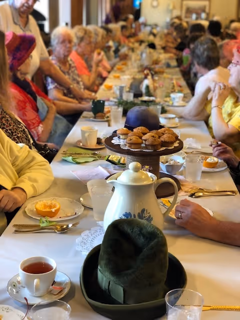 Long dining table in a senior living facility set for tea with a teapot, muffins, and many older adults seated along both sides.
