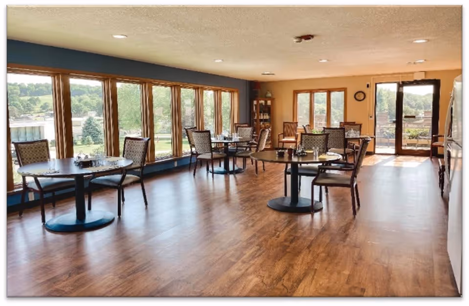 A bright dining room with several round tables and chairs arranged neatly. Large windows line one wall, allowing natural light to fill the space and offering a view of greenery outside. The room has wooden flooring and light-colored walls, with a door leading to an outdoor area.