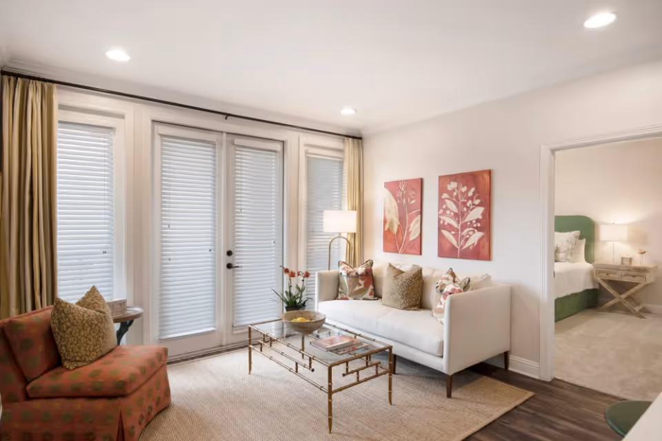 A bright living room with a white sofa adorned with patterned cushions, a glass coffee table with a decorative bowl and books, and a red patterned armchair with a pillow. Behind the sofa are two red floral paintings on the wall. French doors with blinds and beige curtains let in natural light. To the right, an open doorway reveals a bedroom with a green headboard, bedside table, and lamp.