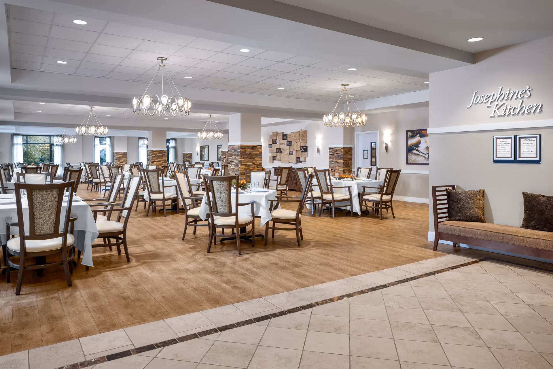 A spacious and well-lit dining room with multiple round tables covered with white tablecloths and surrounded by wooden chairs with cushioned seats. Elegant chandeliers hang from the ceiling, and there is a bench with pillows along the right wall. A sign on the wall reads 'Josephine's Kitchen'.