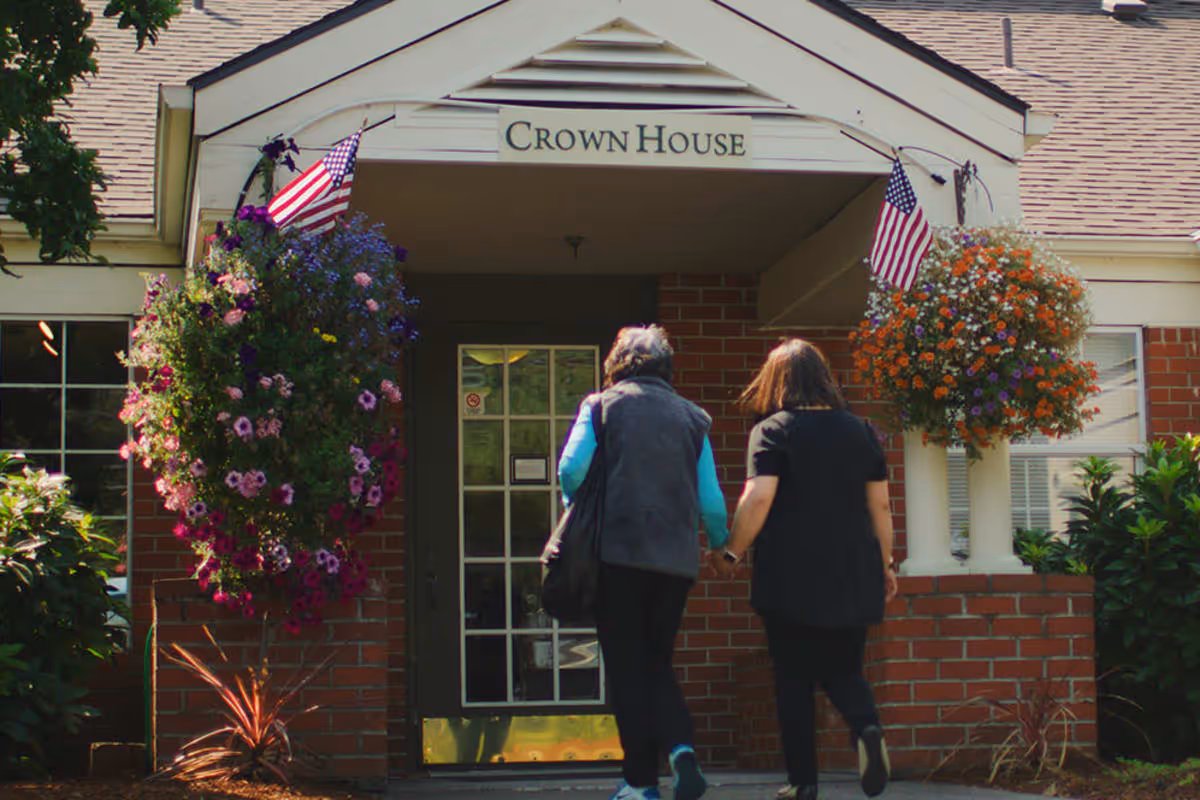 Two women walking hand in hand towards the entrance of a brick building with a sign above the door that reads 'Crown House'. The entrance is decorated with hanging flower baskets and small American flags on either side.