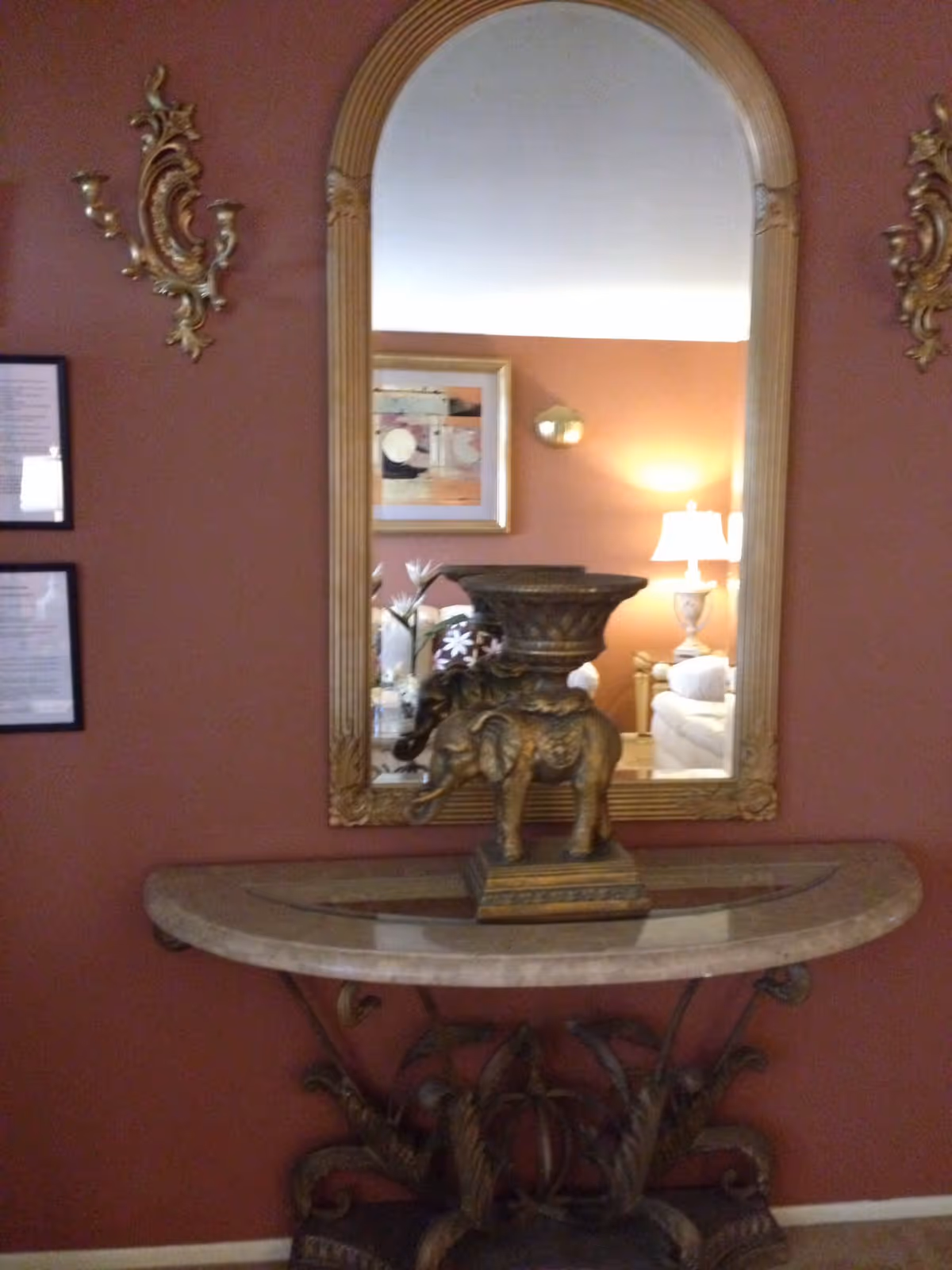 A decorative entryway table with an ornate base and a marble top, holding a golden elephant statue with a bowl on its back. Above the table is a large arched mirror with a gold frame reflecting a living room with a lamp, couch, and wall art. The wall behind the table is painted a warm reddish-brown color and has two gold wall sconces and two framed documents on the left side.