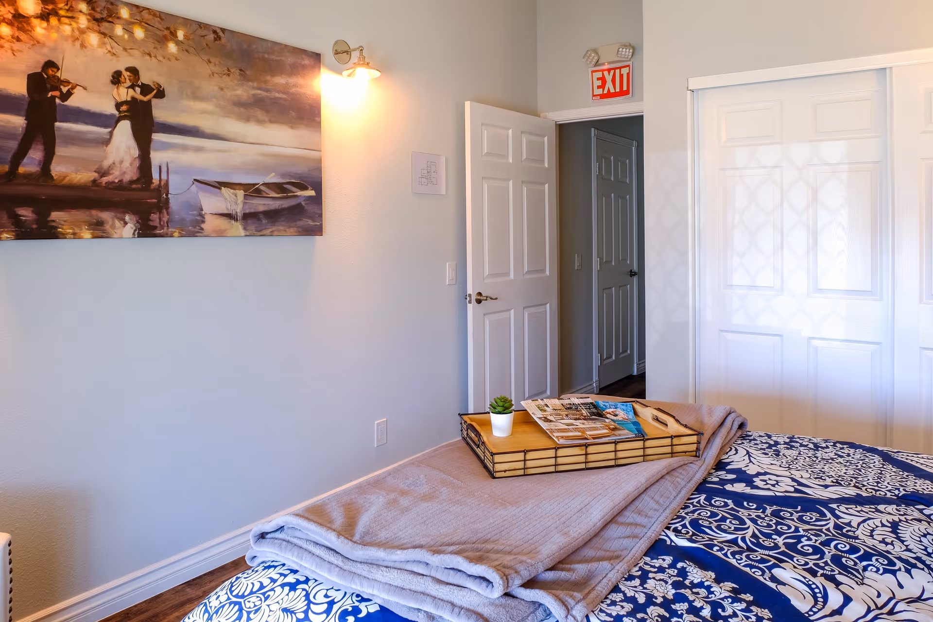 A cozy bedroom with a bed covered in a blue and white patterned bedspread and a folded gray blanket on top. A wooden tray with a small potted plant and magazines rests on the blanket. The room has light-colored walls, a painting of a couple dancing and a violinist on the wall, a wall-mounted light fixture, an open white door leading to a hallway, and a white closet with sliding doors. An illuminated red EXIT sign is above the door.