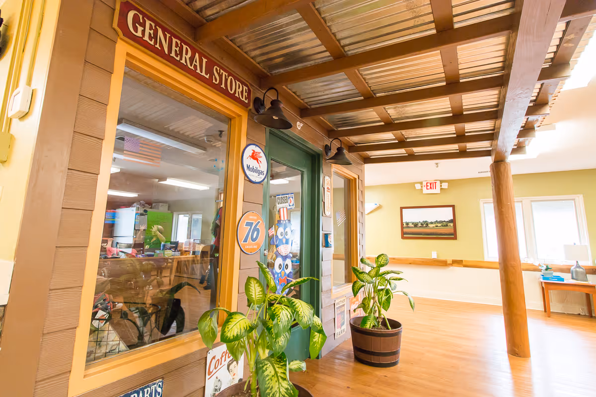 Interior hallway of a senior living facility with a rustic general store facade on the left side, featuring vintage signs and a green door. Two potted plants are placed near the entrance. The hallway has wooden flooring, a wooden beam ceiling, and light yellow walls with a framed landscape painting and an exit sign in the background.