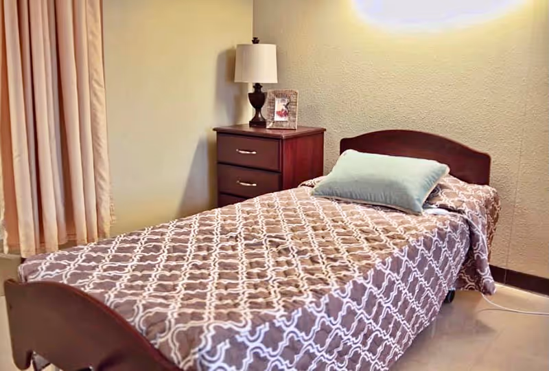 A single bed with a patterned brown and white bedspread and a light blue pillow in a room. Next to the bed is a wooden nightstand with two drawers, a table lamp, and a framed photo. Beige curtains cover a window on the left side of the image.
