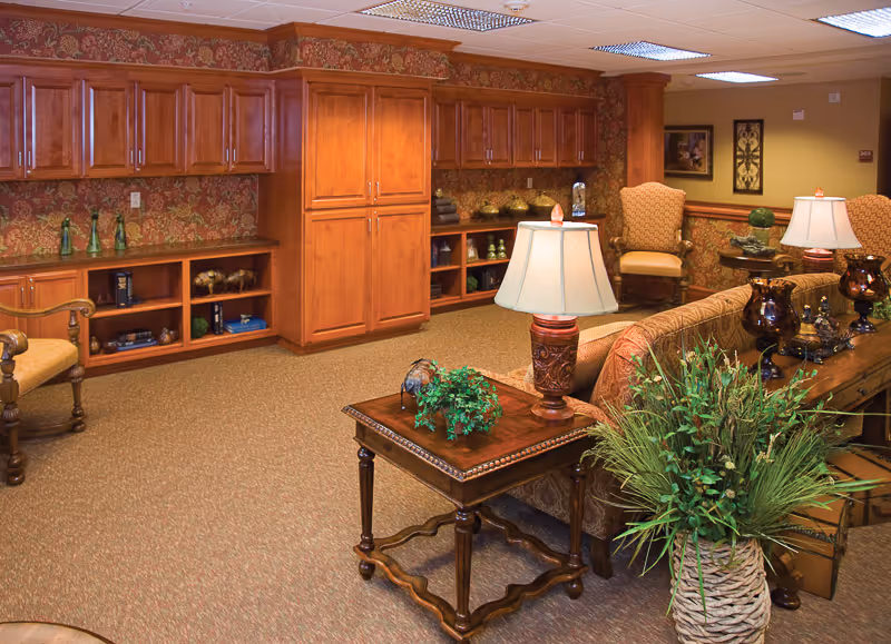 A cozy living room area with wooden cabinetry along the back wall, patterned wallpaper, and carpeted floor. The room features a wooden side table with a lamp and a small plant, a sofa with decorative pillows, two upholstered armchairs, and various decorative items including vases and framed artwork on the walls.