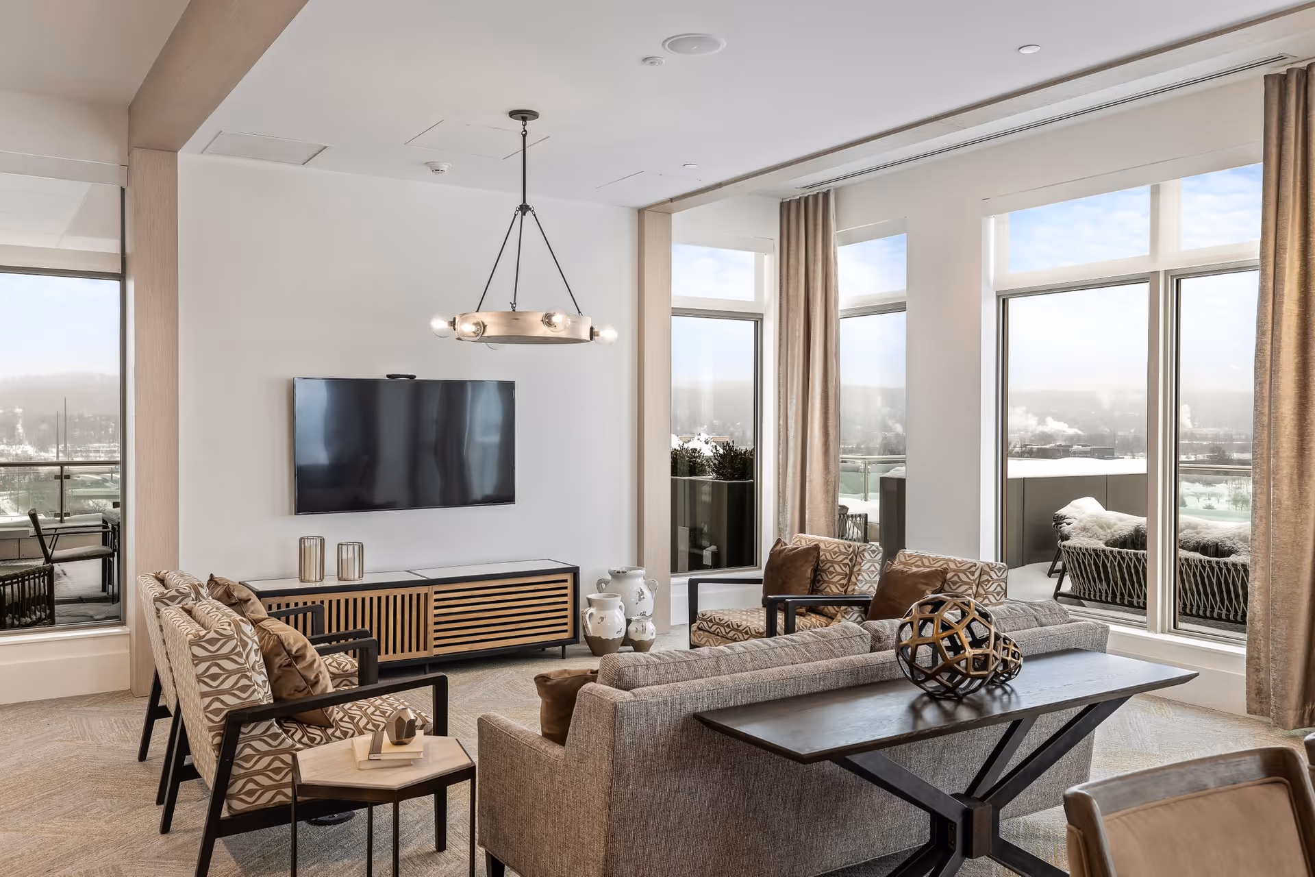 A modern living room with large windows showing an outdoor balcony. The room features a wall-mounted flat-screen TV above a wooden media console, a gray sofa with decorative pillows, two patterned armchairs, a small side table, and a contemporary chandelier hanging from the ceiling. The balcony outside has seating and a view of a snowy landscape.