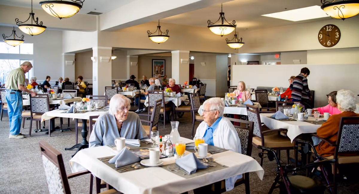 Dining room in a senior living facility with residents seated at multiple tables set for a meal.