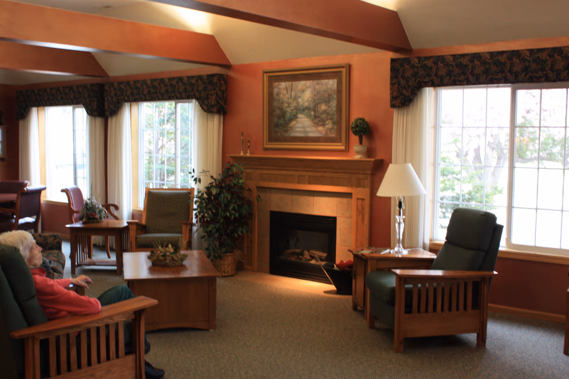 A cozy living room with wooden furniture including armchairs and tables, a fireplace with a painting above it, large windows with curtains letting in natural light, and an elderly person sitting in one of the armchairs.
