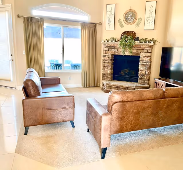 A cozy living room with two brown leather sofas facing a brick fireplace decorated with greenery and framed artwork above. There is a large window with beige curtains and a TV on a stand to the right.
