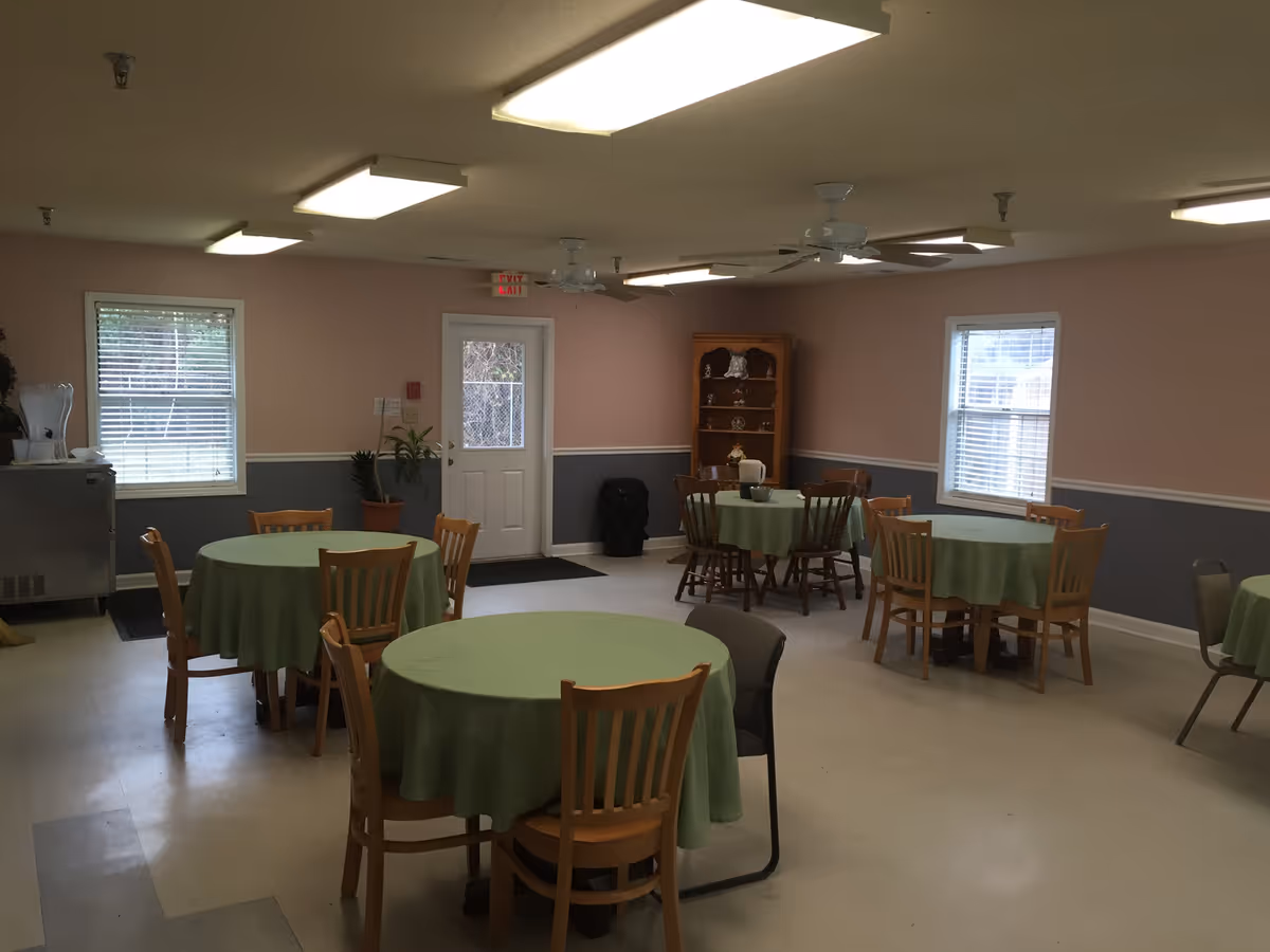 A dining room with several round tables covered with green tablecloths, surrounded by wooden chairs. The room has pink and gray walls, two windows with blinds, ceiling fans, and fluorescent lights. There is a door at the back and a wooden cabinet with decorative items.