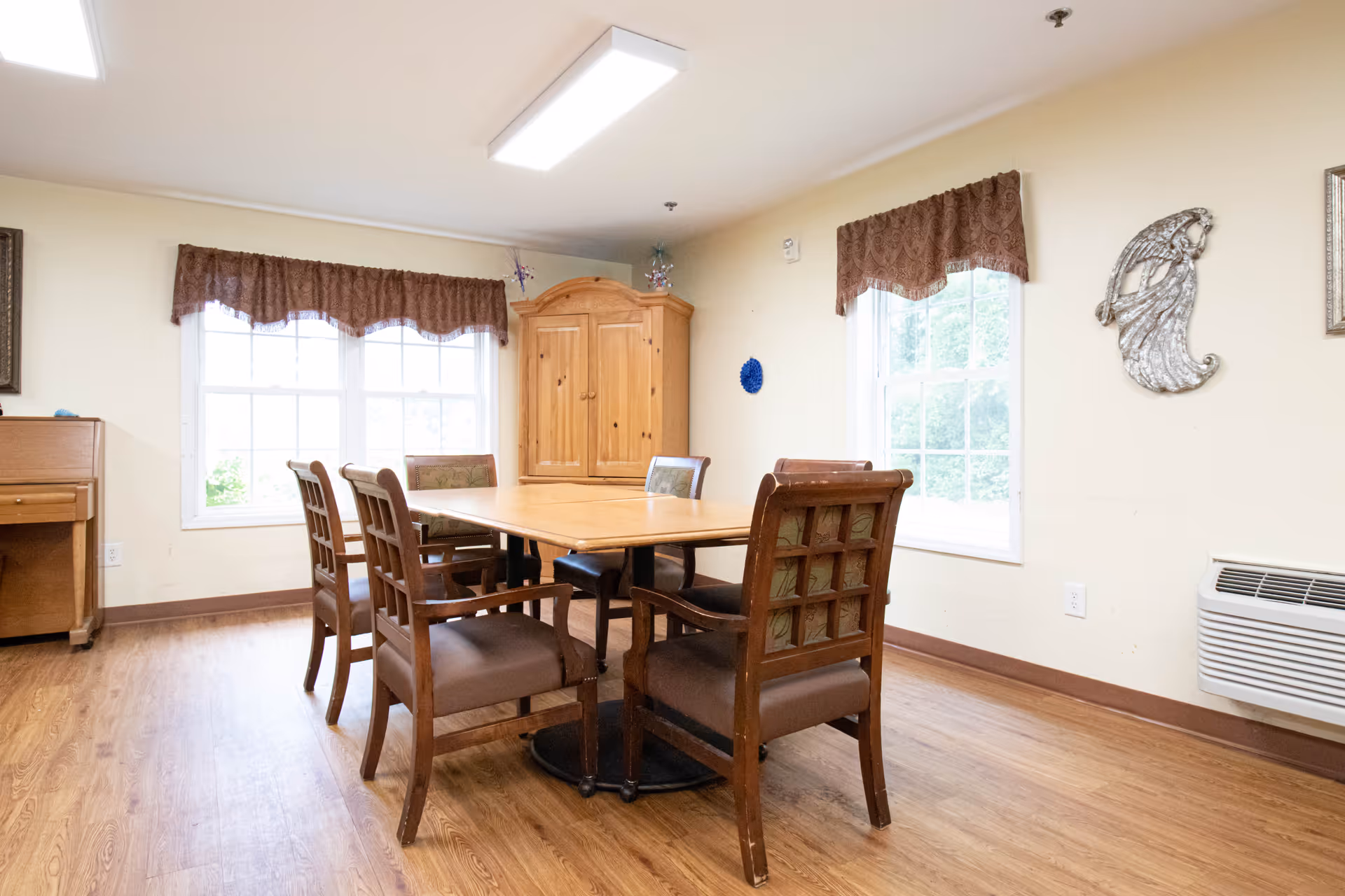 A dining room with a wooden table surrounded by six wooden chairs with cushioned seats. The room has two large windows with brown valance curtains, a wooden cabinet in the corner, a piano on the left side, and a wall-mounted air conditioning unit on the right. The floor is wood, and the walls are light-colored with some decorative wall art.