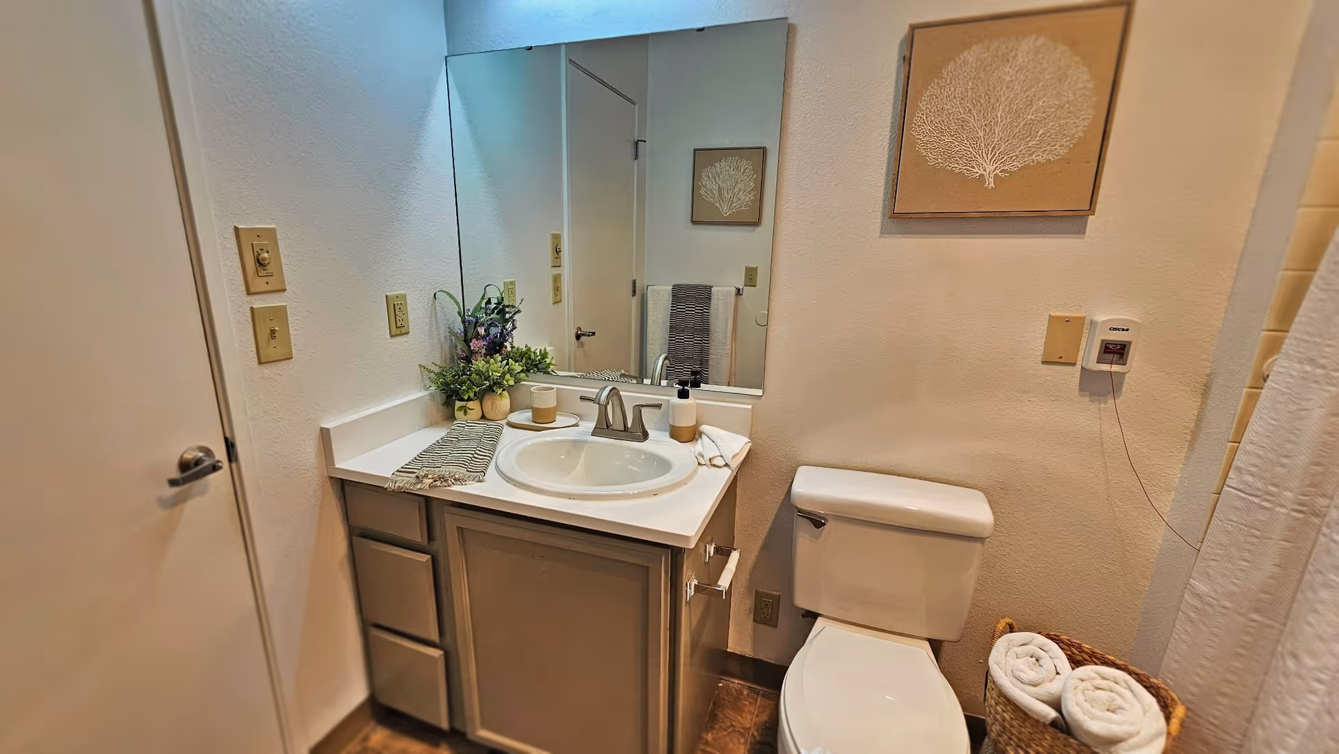 A clean bathroom featuring a white sink with a silver faucet set in a beige countertop, a large mirror above the sink, a white toilet next to the sink, a basket with rolled white towels on the floor, a towel hanging on a rack reflected in the mirror, and a framed coral artwork on the wall.