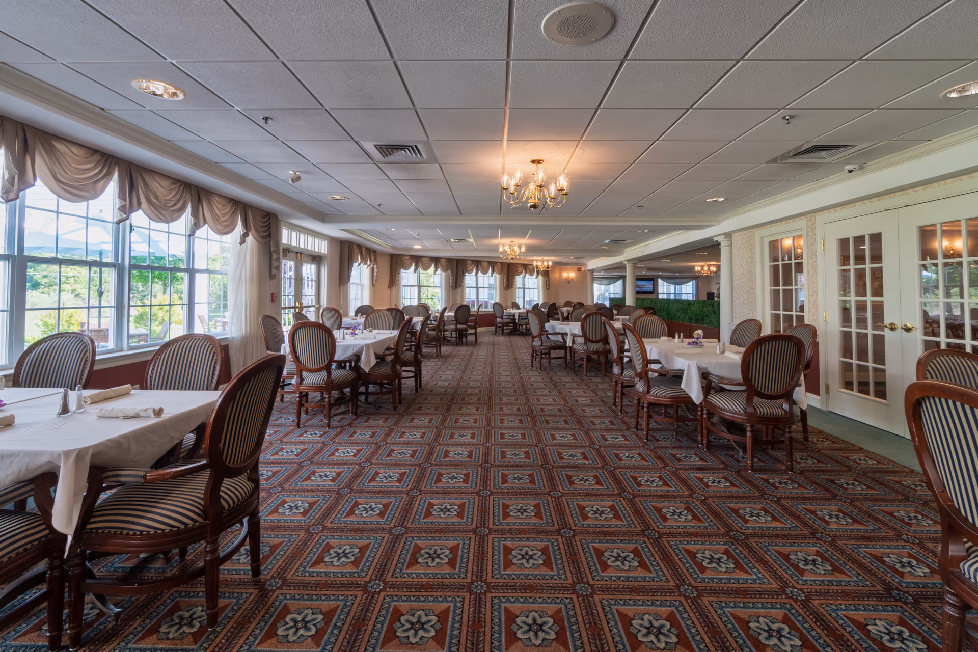 A spacious dining room with multiple tables covered in white tablecloths and set with napkins and silverware. The room features large windows with draped curtains allowing natural light to fill the space. The carpet has a patterned design, and chandeliers hang from the ceiling providing additional lighting. Wooden chairs with striped upholstery surround the tables, and French doors are visible on the right side of the room.