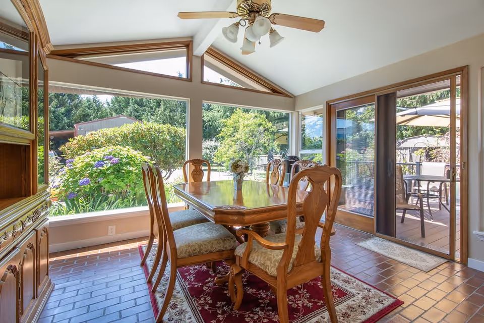 A bright dining room with a wooden dining table and six matching chairs on a red patterned rug. Large windows provide a view of lush greenery outside, and a sliding glass door leads to an outdoor patio with additional seating. A ceiling fan with lights hangs above the table.
