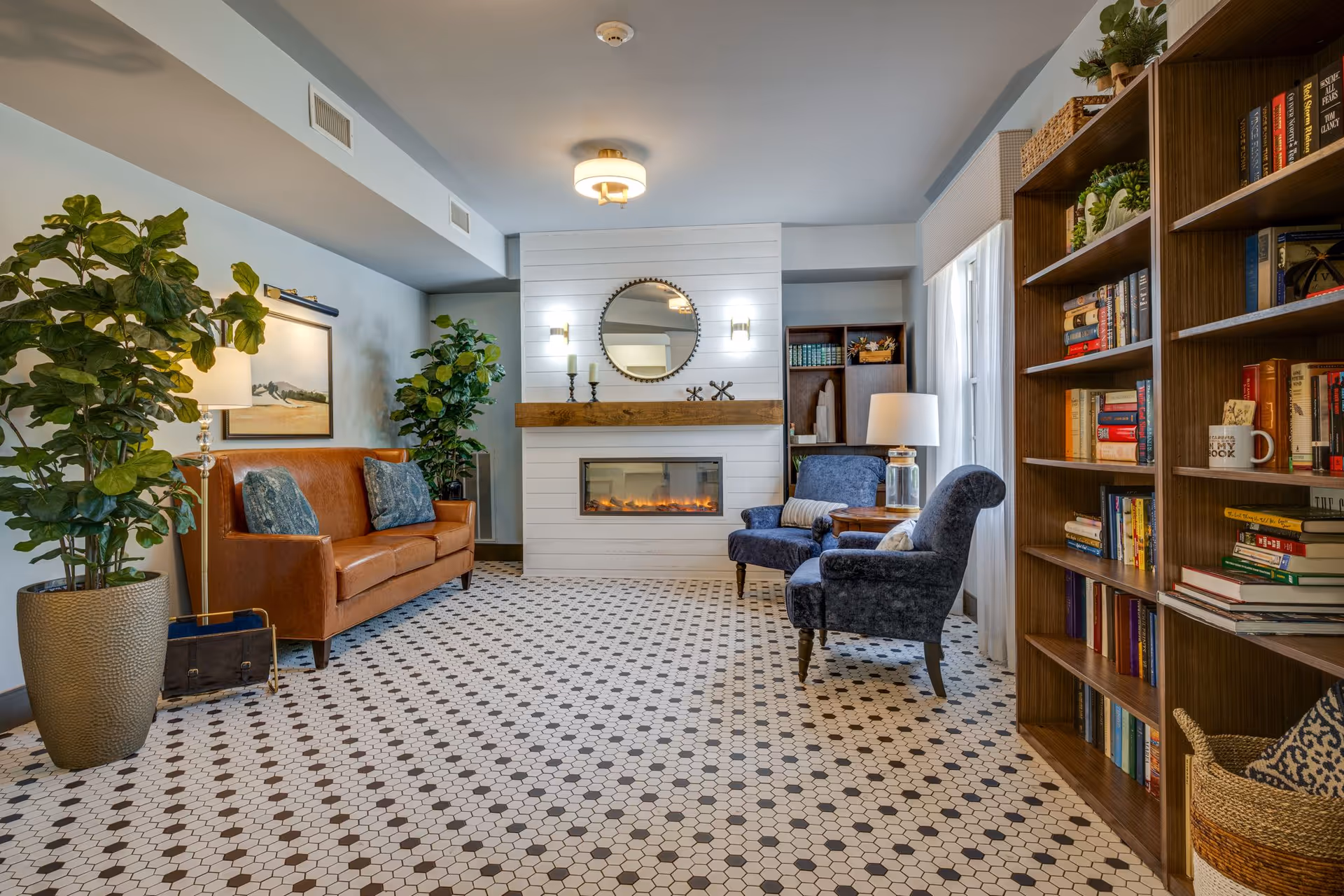A cozy living room with a brown leather sofa adorned with blue pillows on the left, two blue upholstered armchairs with pillows on the right, and a white fireplace with a wooden mantel and a round mirror above it in the center. The floor has a patterned tile design, and there are large potted plants, bookshelves filled with books, and a table lamp near the armchairs. The room is well-lit with natural light coming from a window with sheer curtains on the right.