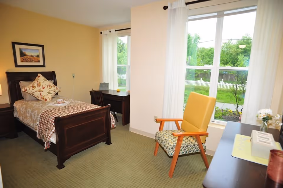 A cozy bedroom in a senior living facility with a single wooden bed adorned with patterned bedding and pillows. There is a wooden nightstand with a lamp beside the bed, a wooden desk with a chair near the window, and a cushioned armchair with wooden arms positioned by a large window with white curtains, overlooking a green outdoor area with a white picket fence.