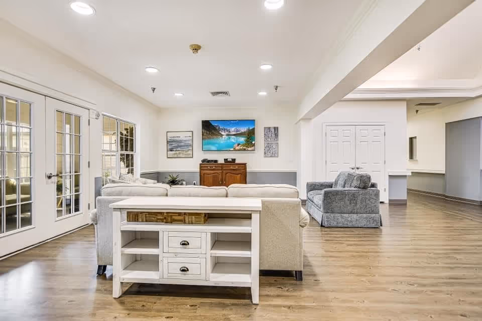 A spacious and well-lit senior living facility common area featuring light-colored walls and wood flooring. The room includes a beige sofa with a white console table behind it, a gray armchair, and a wooden cabinet against the far wall. Above the cabinet is a flat-screen TV displaying a scenic mountain and lake image, flanked by framed artwork. Large glass doors on the left side allow natural light to enter the room.