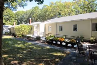 Single-story beige Olsen Rest Home exterior with ramp, lawn, and flowering planters.