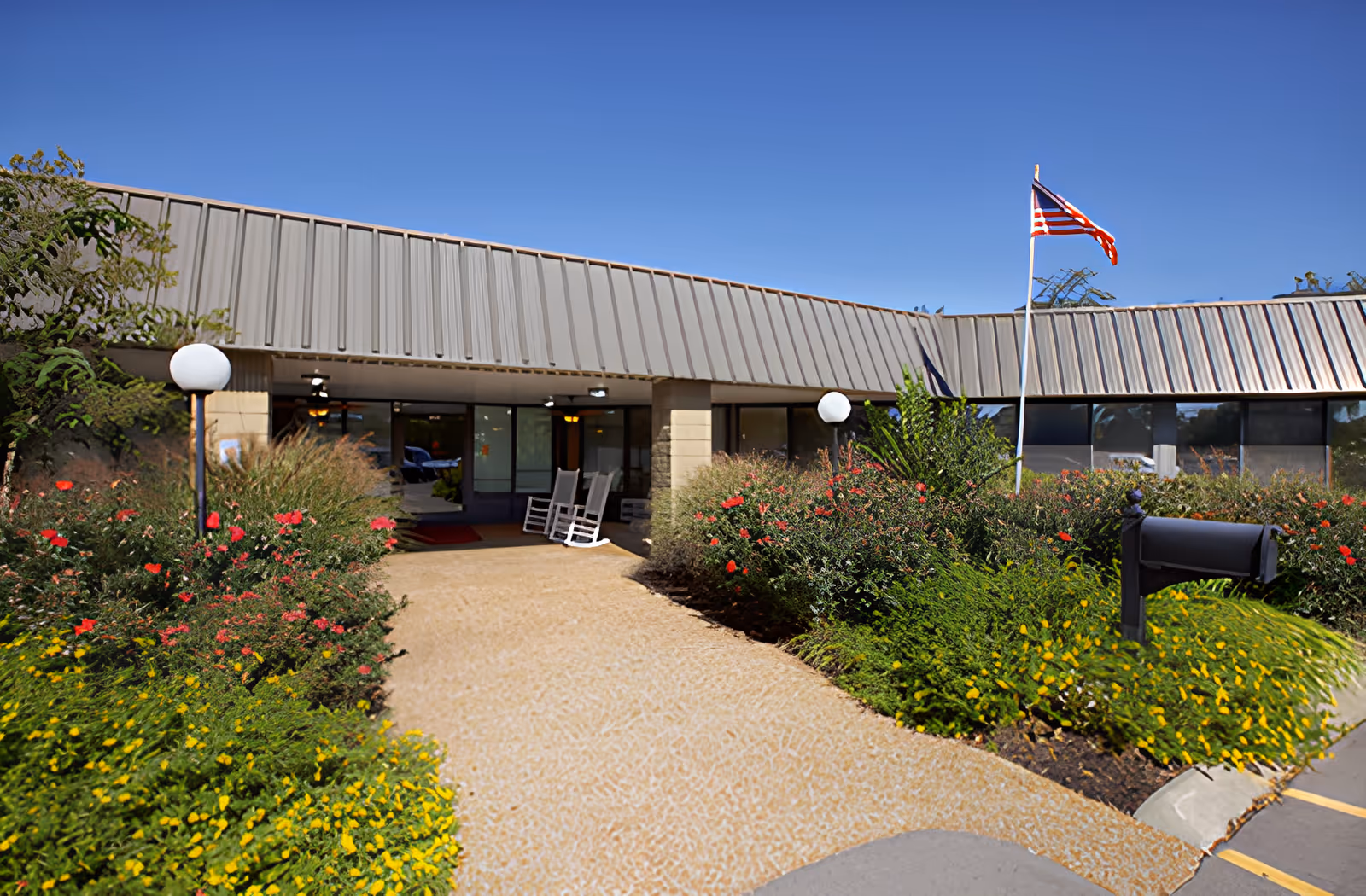 Entrance to a single-story building with a metal roof, surrounded by well-maintained flower beds with yellow and red flowers. There are two white rocking chairs near the glass doors at the entrance, an American flag on a pole to the right, and a black mailbox in the foreground. The sky is clear and blue.