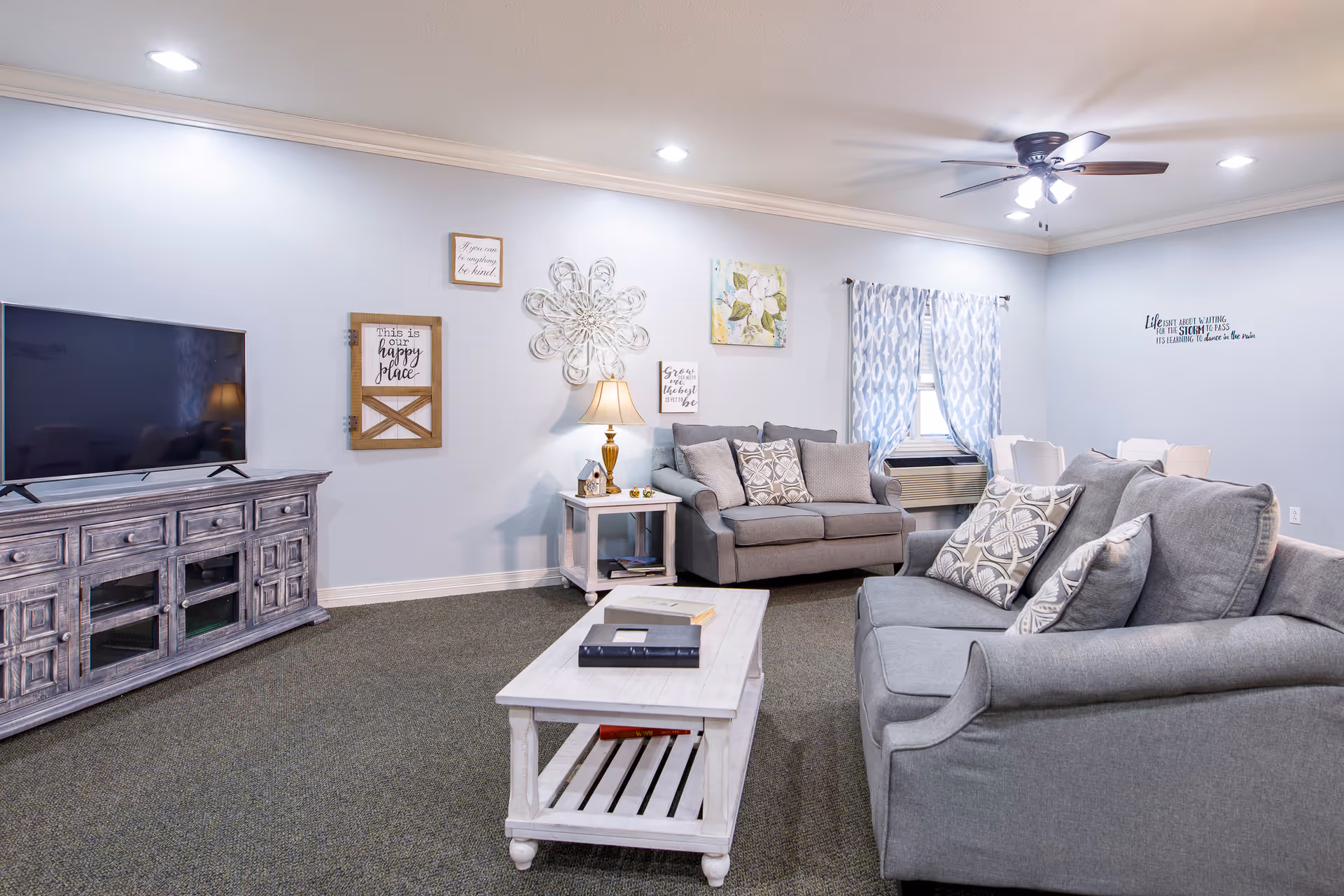 Bright furnished living room with gray sofas, a TV on a wooden console, a coffee table, wall decor, and a ceiling fan.