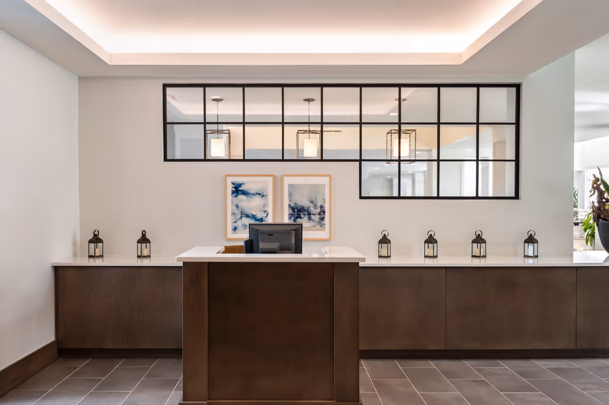 A modern reception desk area with a dark wood front desk and a long counter behind it. The wall behind the desk features a large window with black grid framing, two abstract blue and white paintings, and six decorative lanterns evenly spaced on the counter. The ceiling has recessed lighting with a soft glow.