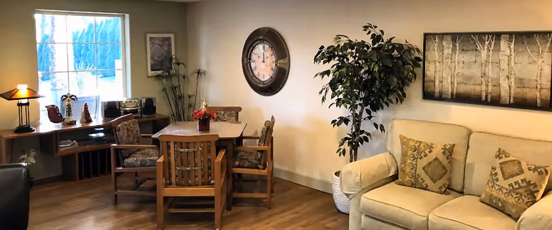 A cozy interior room with a wooden table and four chairs, a beige sofa with patterned cushions, a large wall clock, a potted plant, and a side table with decorative items and a lamp near a window.