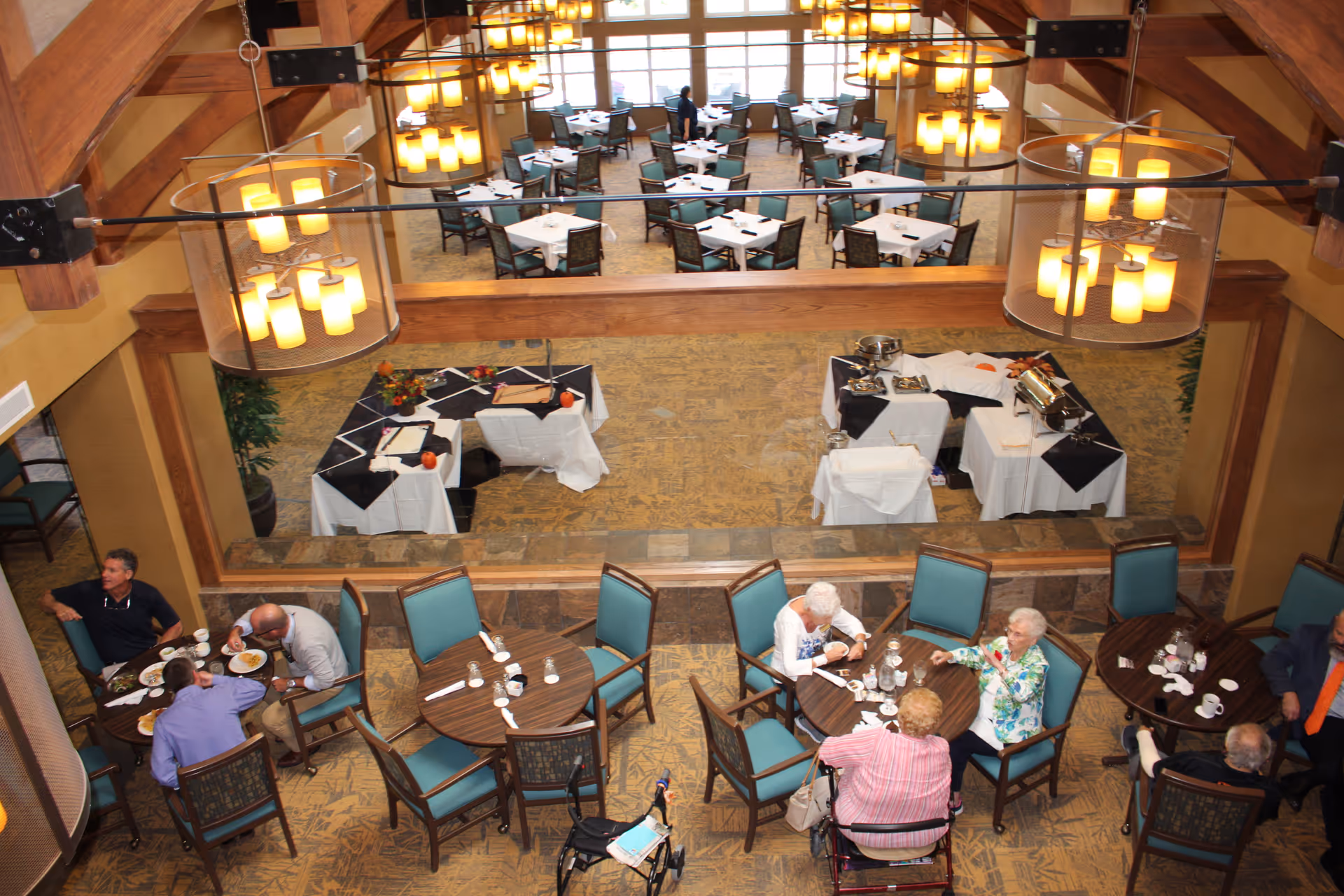 Overhead view of a dining area in a retirement community with several round tables and teal cushioned chairs. Elderly residents are seated at some tables eating and conversing. The room features warm lighting with hanging chandeliers and a large mirror reflecting additional tables and chairs set with white tablecloths and black overlays.
