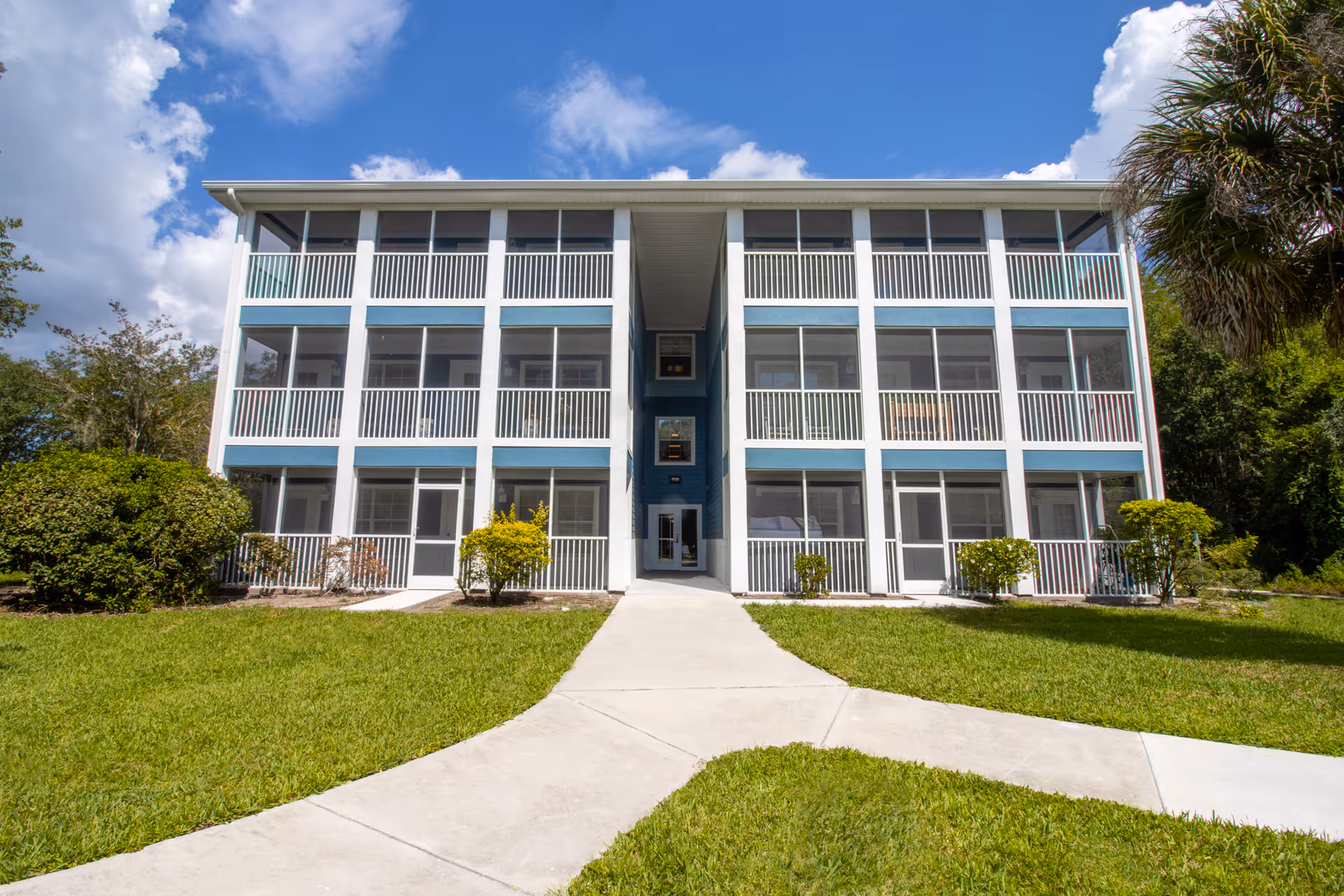 Front exterior view of a three-story residential building with screened balconies on each floor, surrounded by green grass, bushes, and trees under a partly cloudy blue sky.