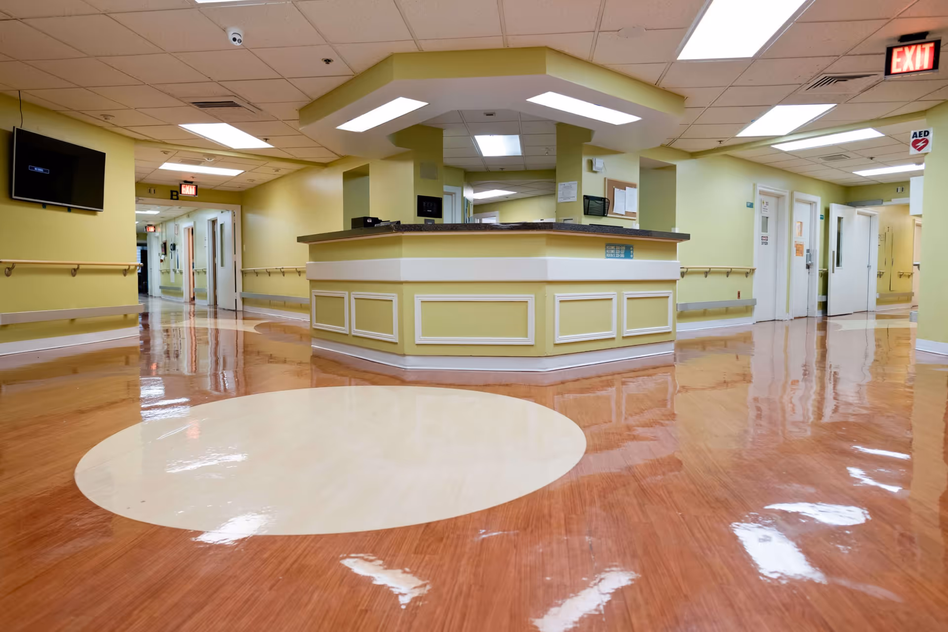 Interior view of a healthcare facility hallway with a central nurse's station. The walls are painted light yellow, and the floor has a polished wood finish with a large light-colored circular pattern. Handrails line the walls, and there are multiple doors and exit signs visible. A television is mounted on the left wall, and an AED sign is visible on the right.