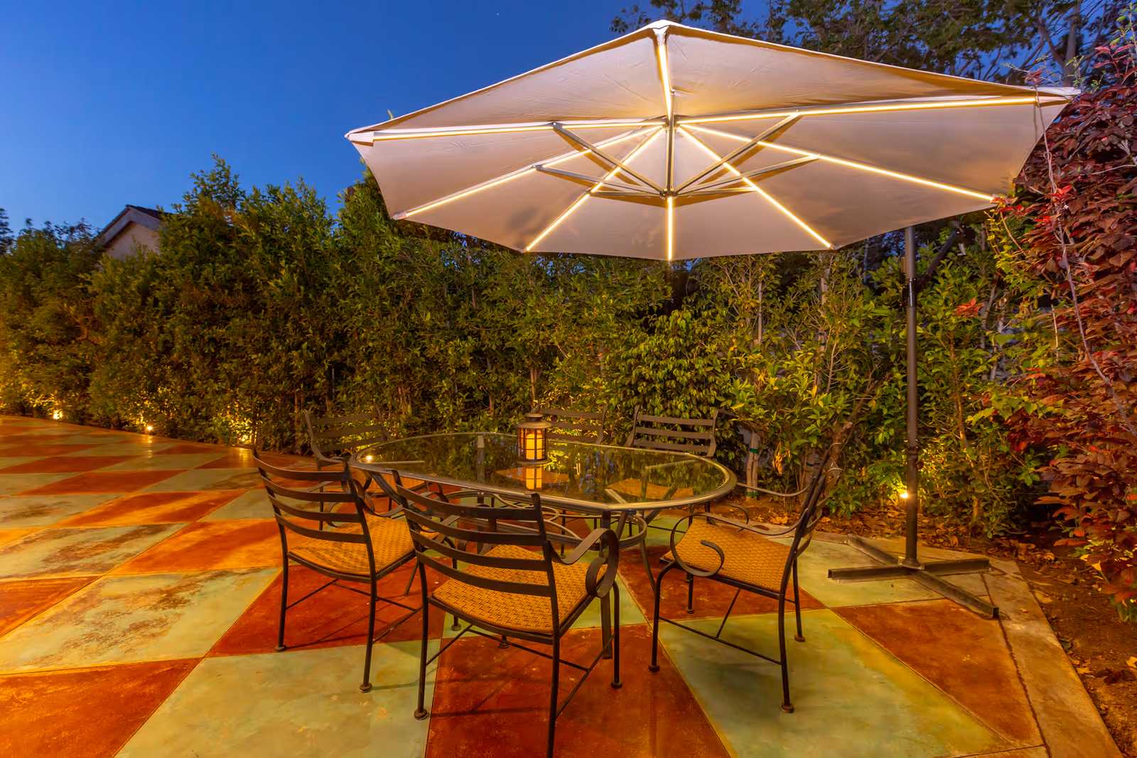 Outdoor patio at dusk with a glass-top table, metal chairs, and a large illuminated umbrella on a patterned tiled courtyard surrounded by hedges.