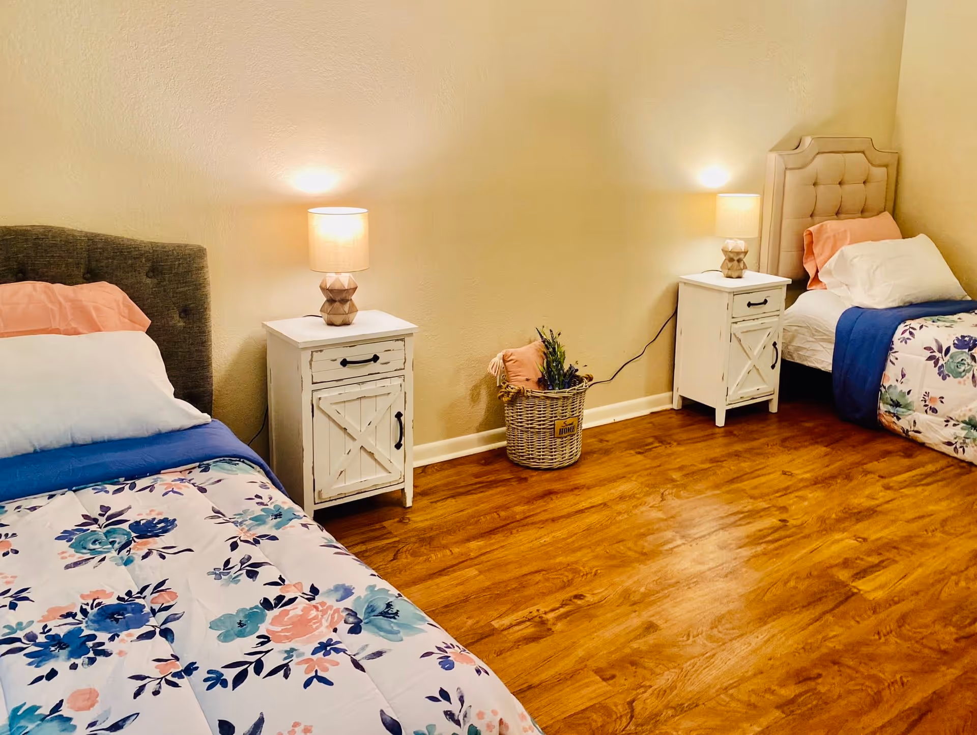Shared bedroom with two twin beds, floral bedding, nightstands with lamps, and a hardwood floor.