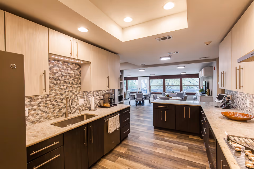 Modern kitchen area with light and dark wood cabinets, a tiled backsplash, a sink, and countertop appliances. The kitchen opens into a spacious dining and living area with large windows letting in natural light.