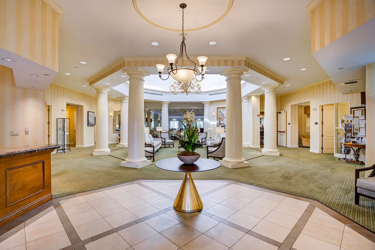 Spacious and elegant senior living facility lobby with a round table holding a plant in the center, surrounded by white columns and comfortable seating areas. The room features a chandelier, carpeted floors, and soft yellow striped walls.