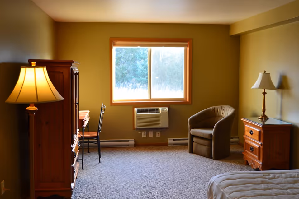 A cozy senior living bedroom with a window showing outdoor greenery. The room features a wooden dresser with a lamp, a small desk with a chair, a cushioned armchair, a nightstand with a lamp, and part of a bed with white bedding. The walls are painted a warm beige color and the floor is carpeted.