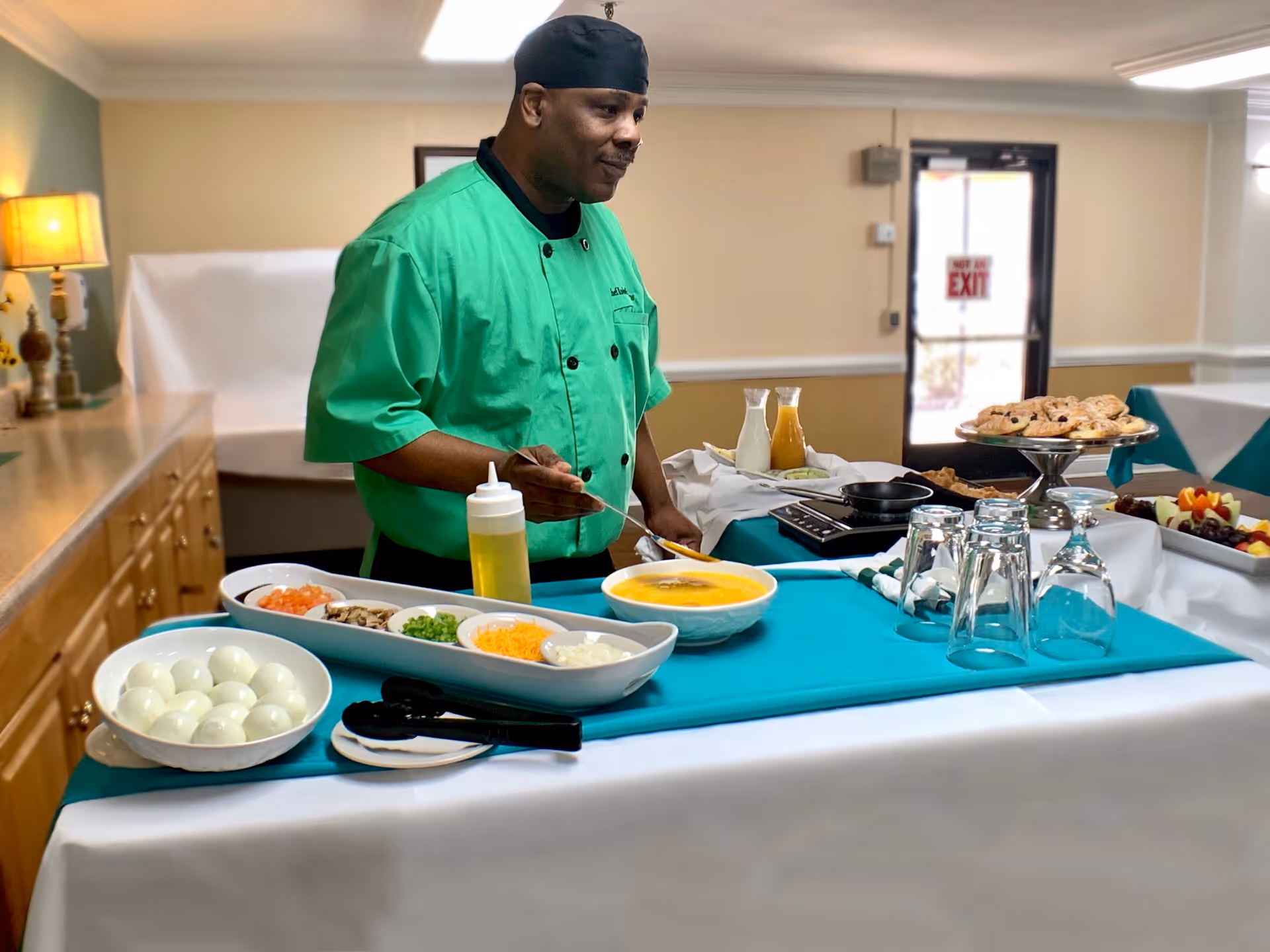 A chef wearing a green uniform and black hat stands behind a table set with various food items including a bowl of soup, a plate of boiled eggs, chopped vegetables, shredded cheese, and condiments. The table is covered with a white and teal tablecloth, and there are glasses and plates arranged on it. The background shows a room with beige walls, a lamp on a wooden cabinet, and a door with a 'Not an Exit' sign.