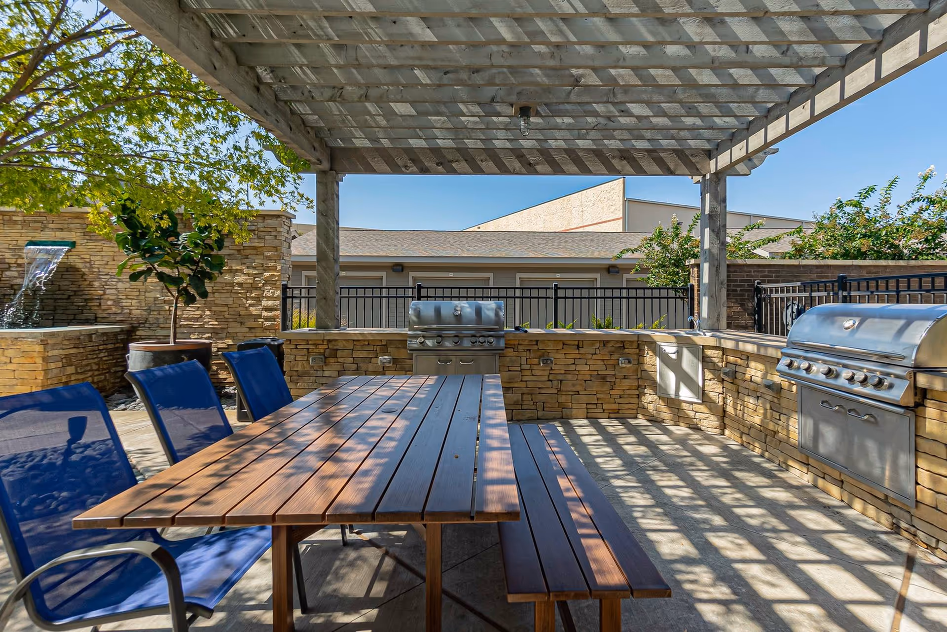 Outdoor patio area with a wooden pergola overhead casting shadows on a wooden picnic table and bench with blue chairs. There are two stainless steel grills built into a stone counter, a small sink, and a water feature with a waterfall on the left side. Trees and a building are visible in the background under a clear blue sky.